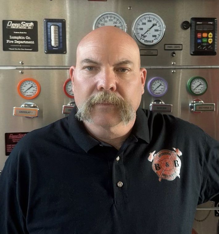 A bald man with a beard and mustache is standing in front of a display of gauges