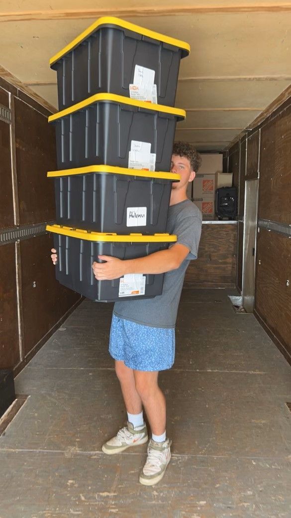 A man is carrying a stack of black plastic bins with yellow lids.