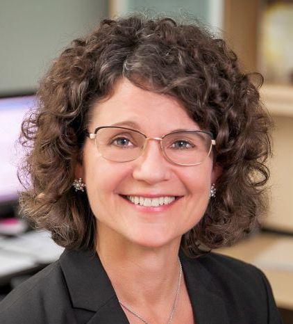 Woman with curly brown hair, glasses, smiling, wearing a black blazer.