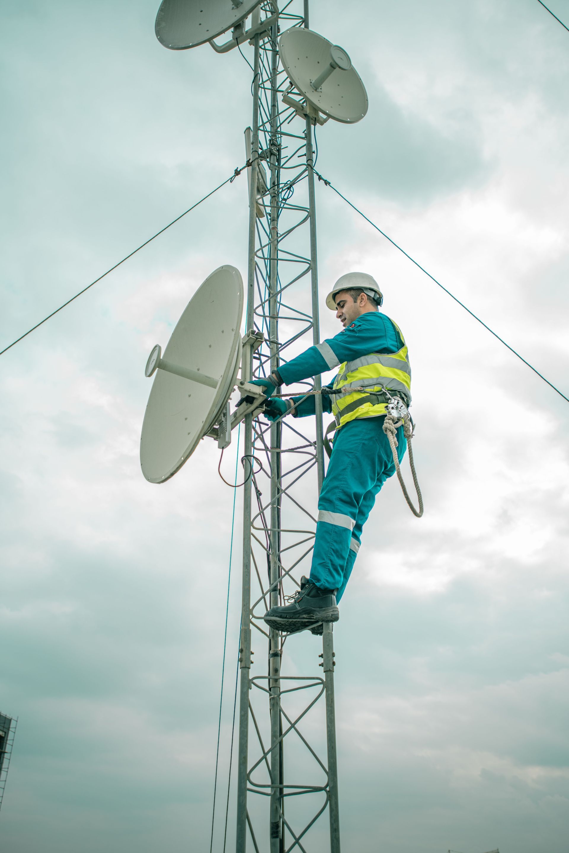 Worker in blue safety gear climbs a tall communications tower with satellite dishes.