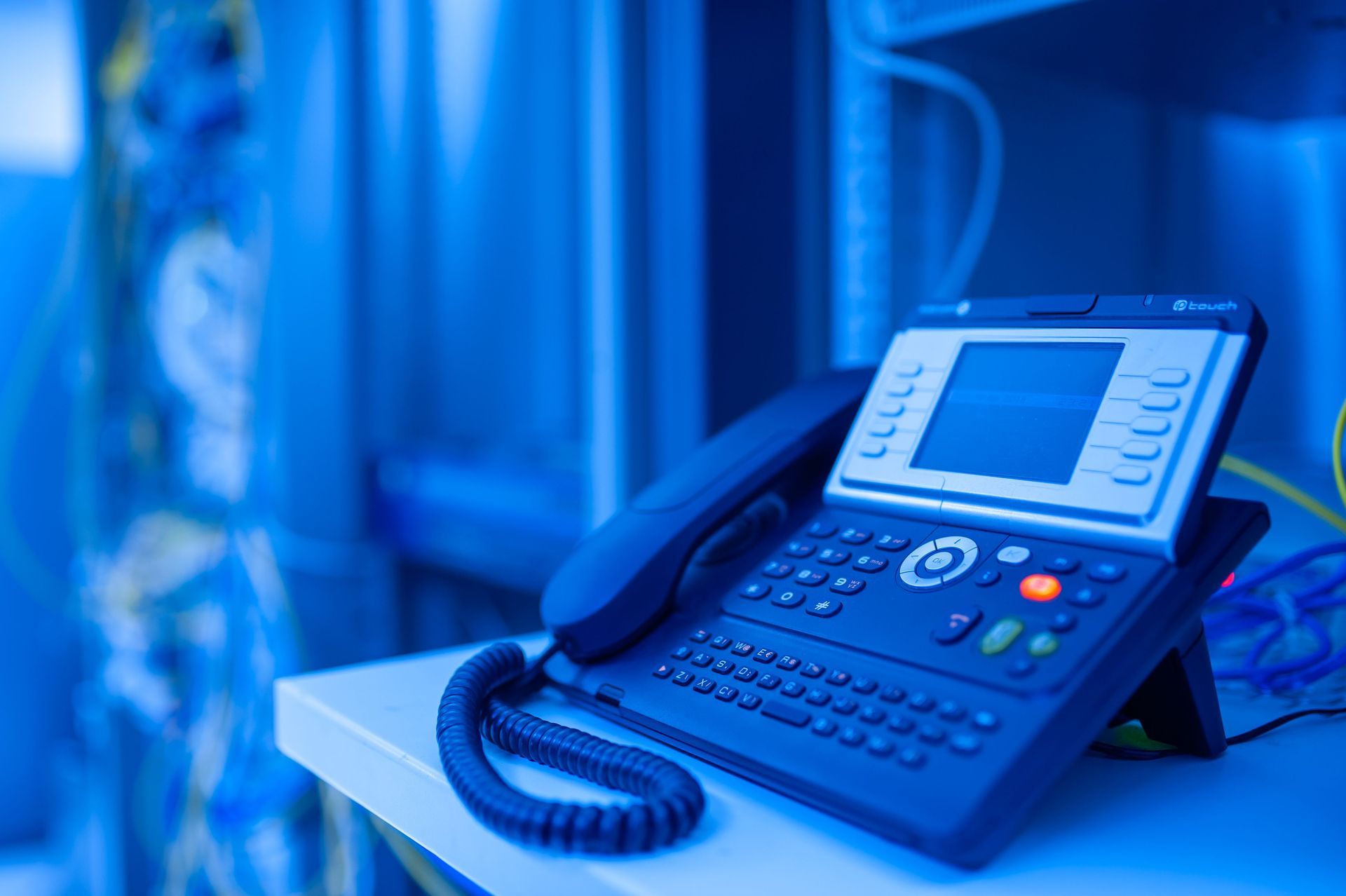 Blue office desk phone with keypad and handset on a desk in a blue-lit office