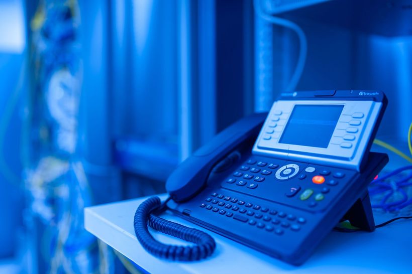 Blue office desk phone with keypad and handset on a desk in a blue-lit office