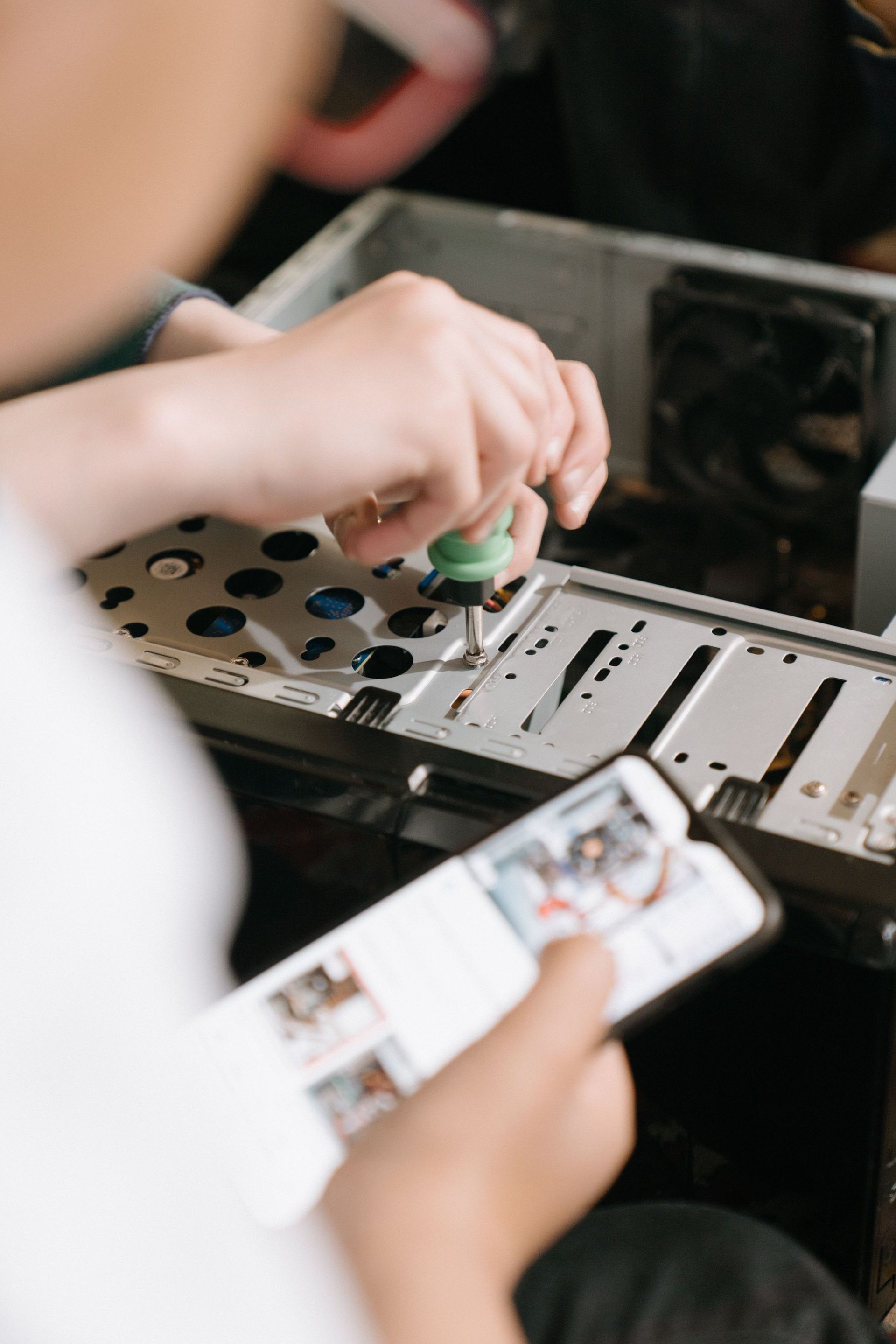 Hands using a screwdriver to repair a computer tower beside a smartphone with instructions on screen