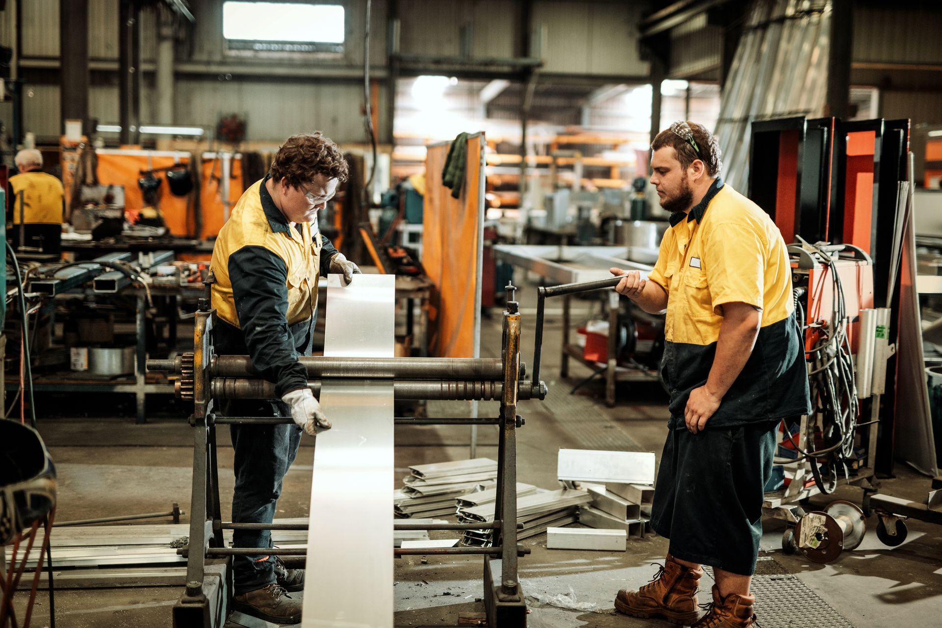 Two metal fabricators in yellow shirts operating machinery in a factory with safety gear 