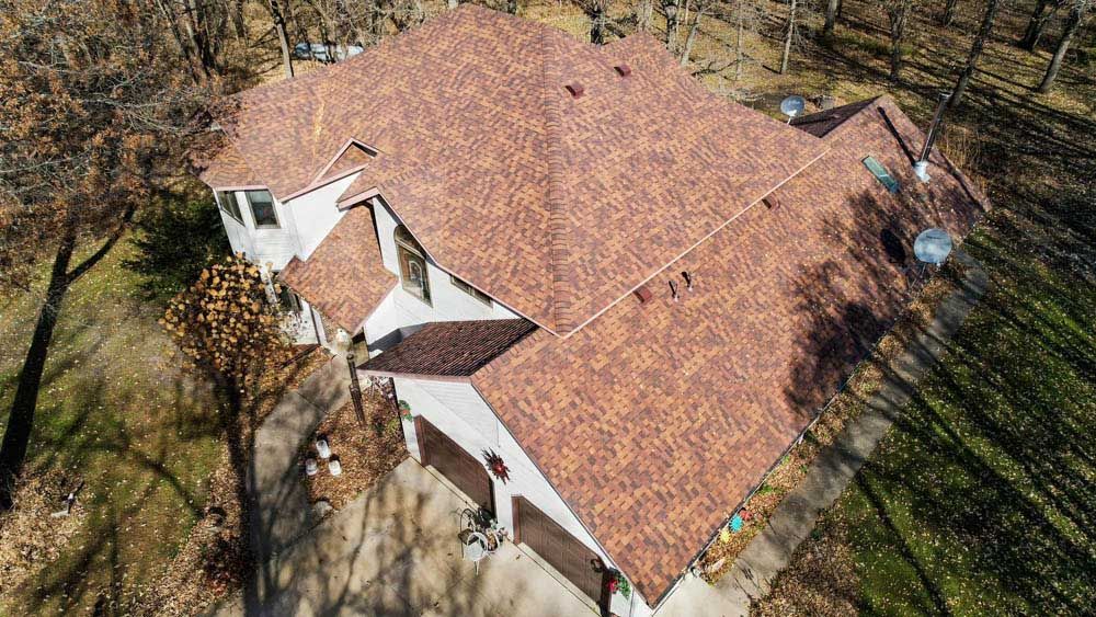 Aerial view of a tan house with a reddish-brown roof surrounded by trees and a driveway.