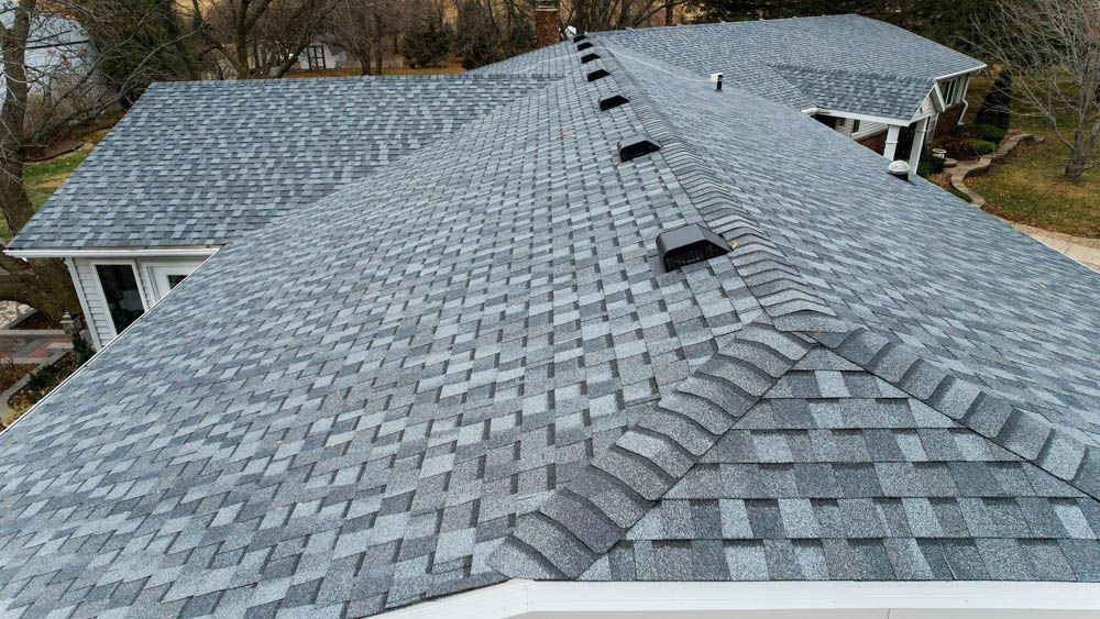 Aerial view of a gray shingled roof with multiple dormers and a chimney in a wooded area