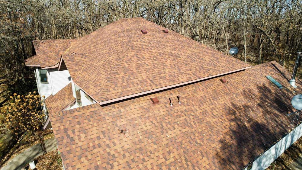Aerial view of a large tan shingle roof surrounded by leafless trees.