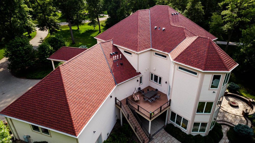 Aerial view of a large white house with a red roof and wraparound deck, surrounded by green trees.