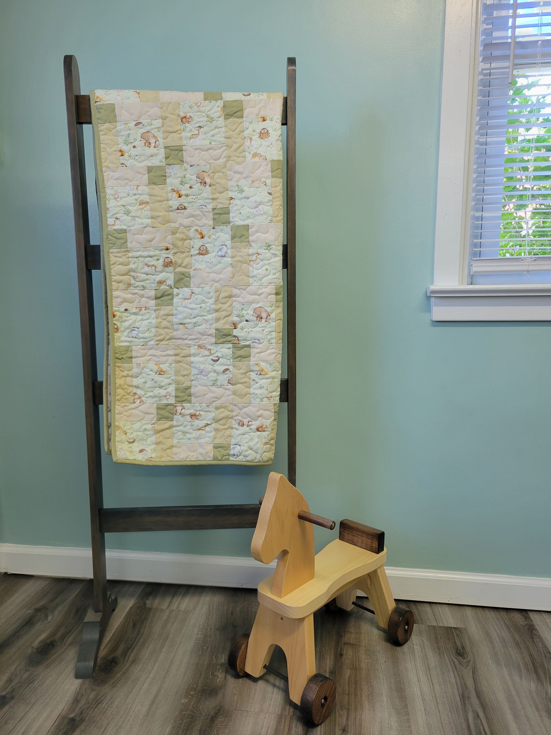 Quilt on a wooden frame beside a small rocking horse in a light blue room.