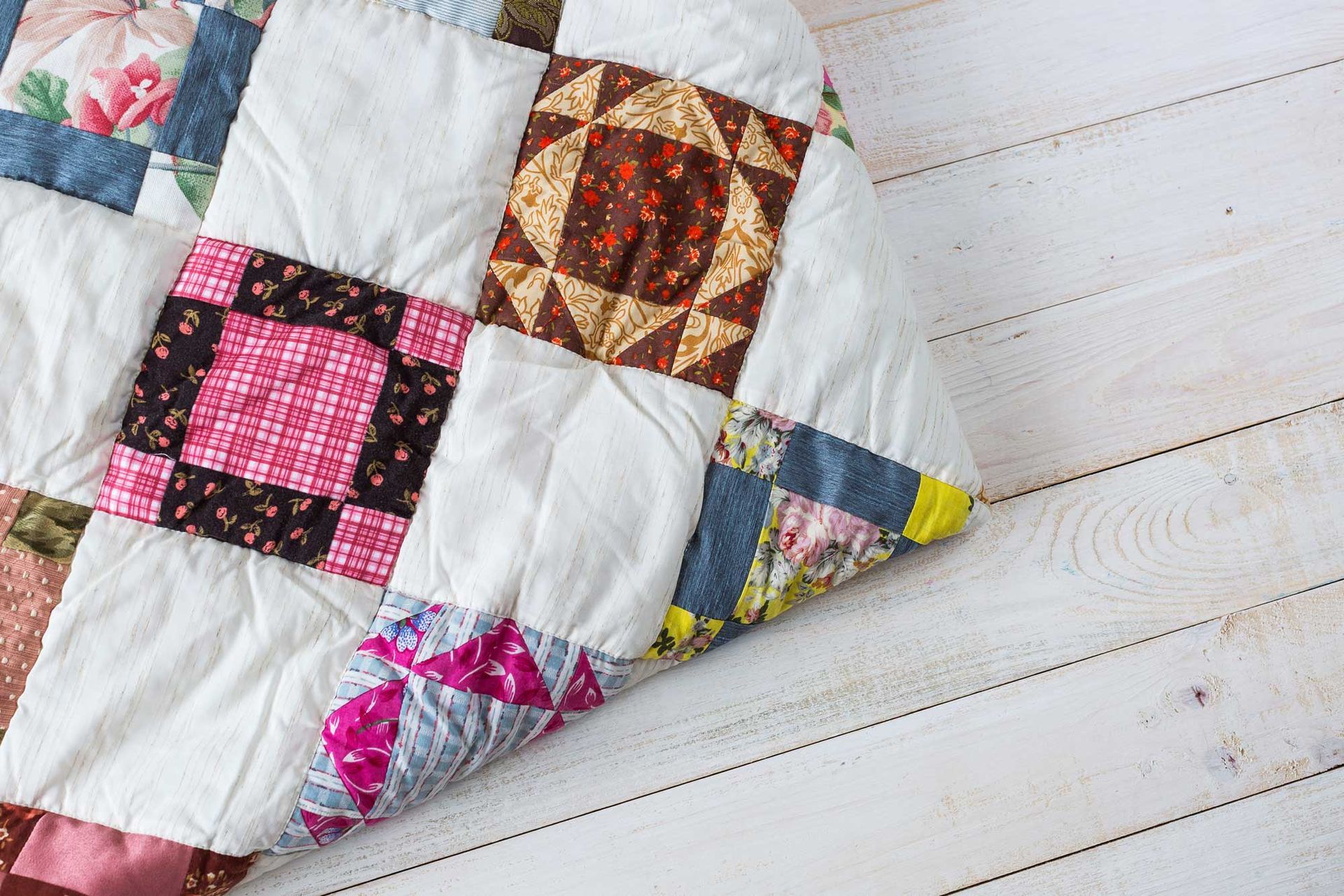 A folded patchwork quilt with colorful patterned fabric squares on a light wooden floor. A folded patchwork quilt with colorful patterned fabric squares on a light wooden floor.
