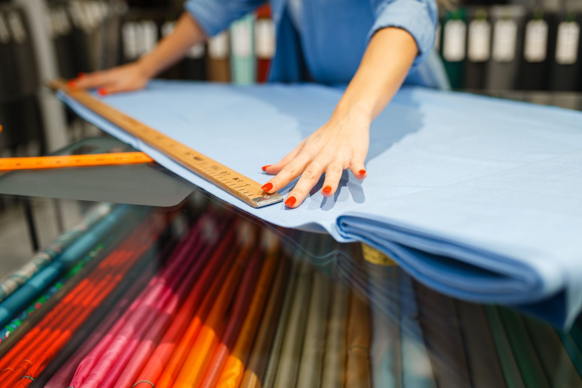 Hands measuring folded blue fabric with a ruler on a table, surrounded by colorful fabric rolls. Hands measuring folded blue fabric with a ruler on a table, surrounded by colorful fabric rolls.