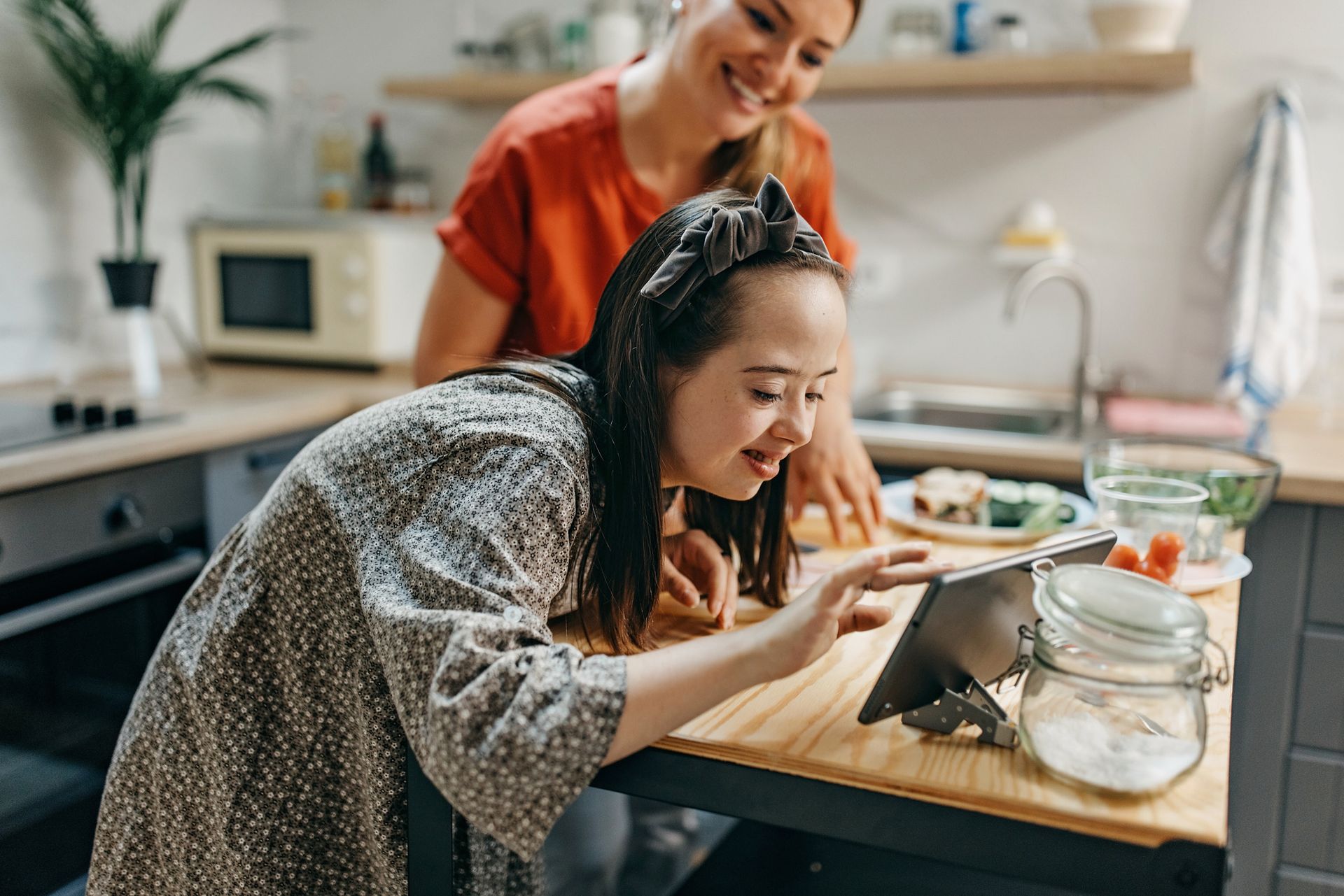 Woman and girl in kitchen looking at tablet, smiling.