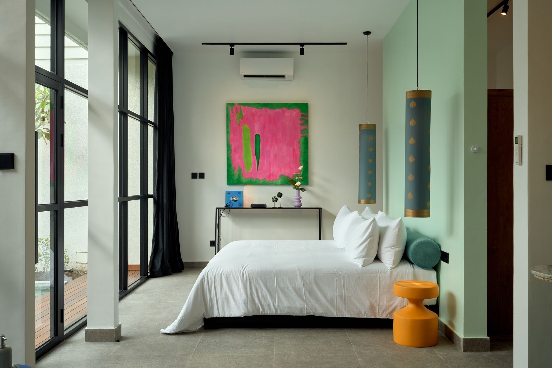 Bright modern bedroom with white bed, pink wall art, large windows, and orange stool