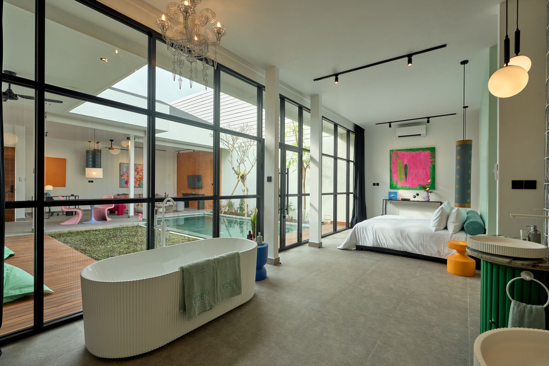 Modern bedroom with glass walls, white bed, green rug, and colorful wall art facing a courtyard
