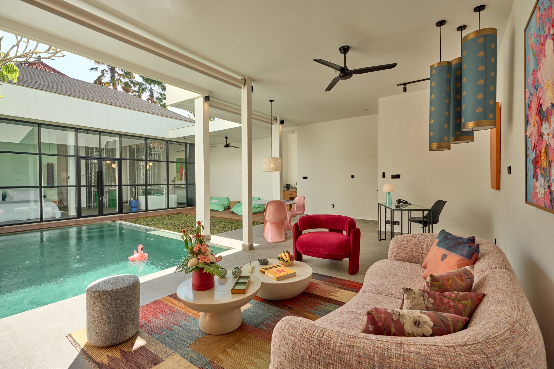 Bright living room opening to a pool, with a person seated on a red chair and a pink sofa in the foreground.