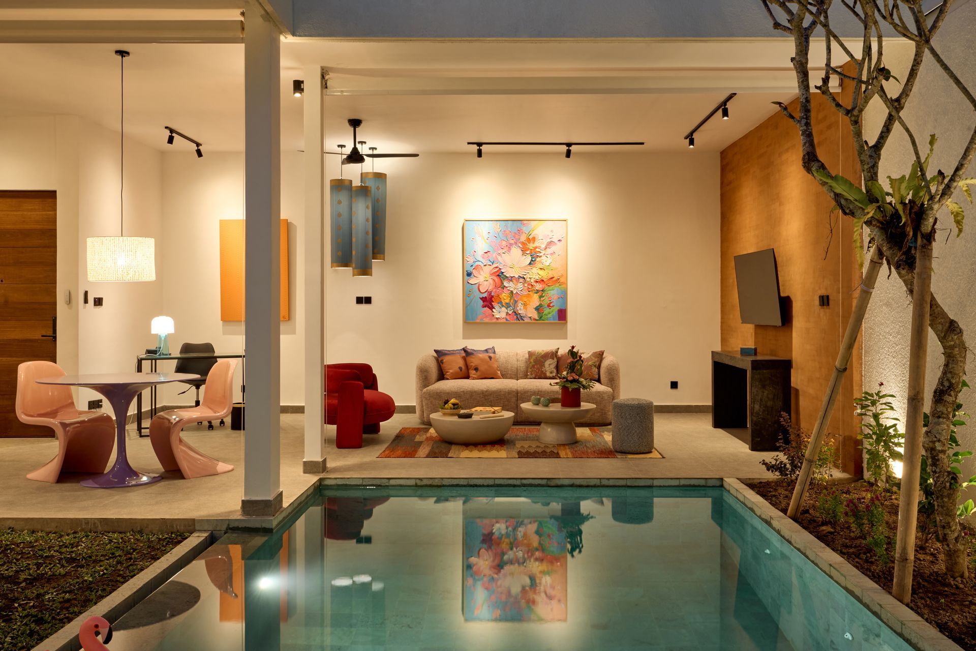Modern living room with warm lighting and a pool in the foreground, featuring colorful wall art and red chairs