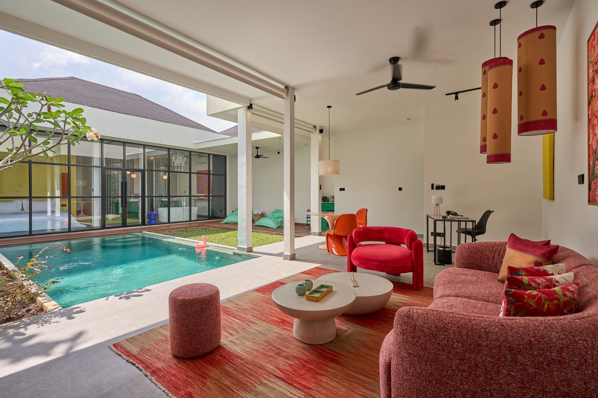 Bright living room opening to a pool, with a person seated on a red chair and a pink sofa in the foreground.