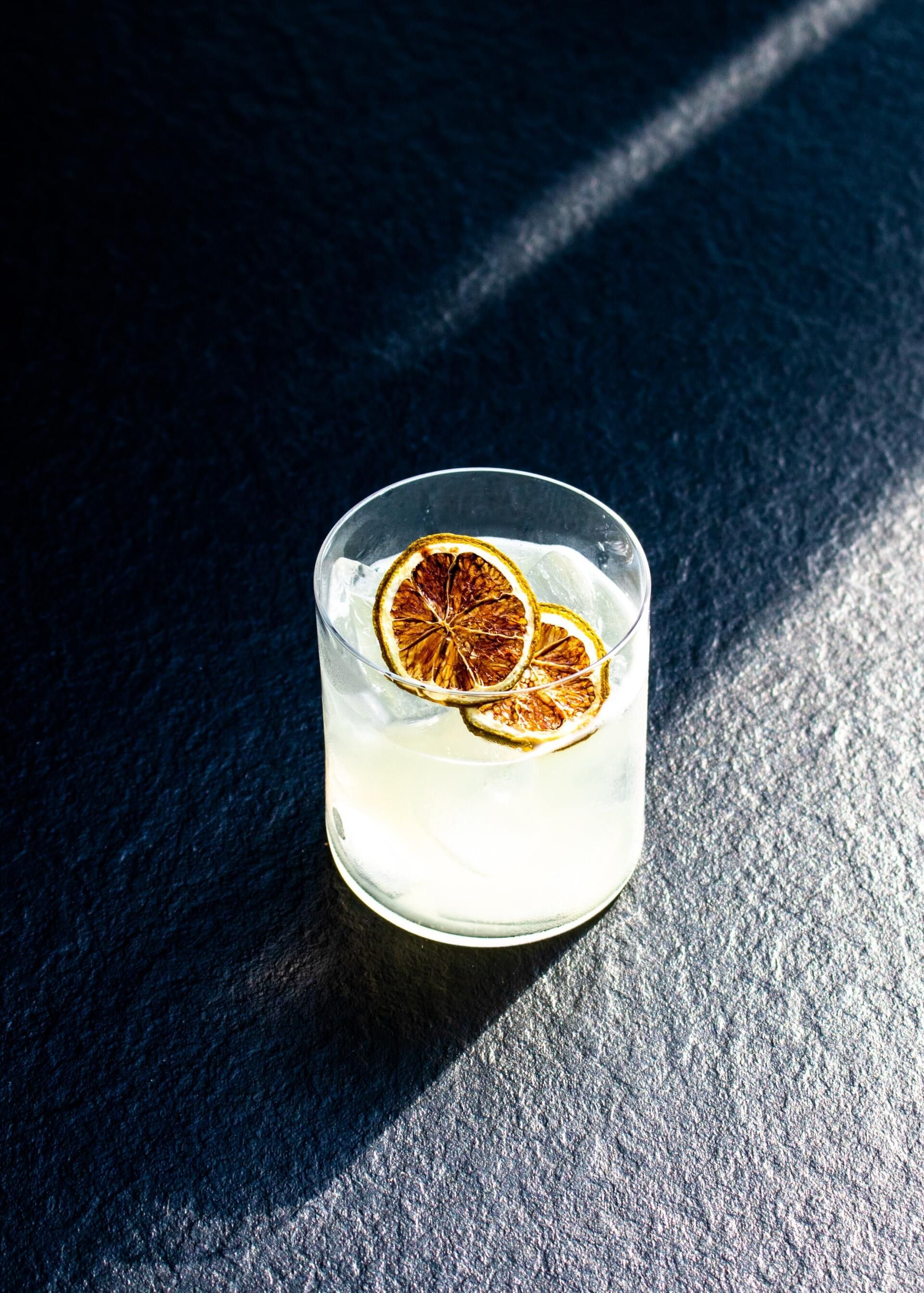 Clear drink in a glass with dried citrus slices on a dark textured surface, lit by sunlight.