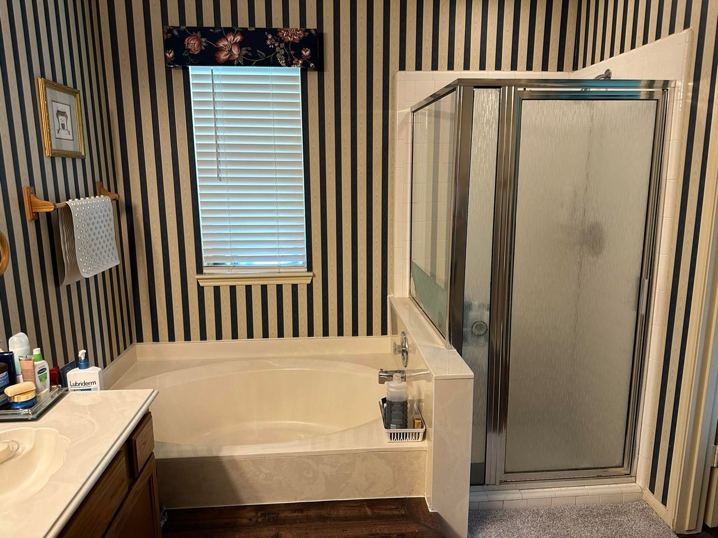 Small bathroom with striped wallpaper, bathtub, glass shower, and vanity under a window with blinds.