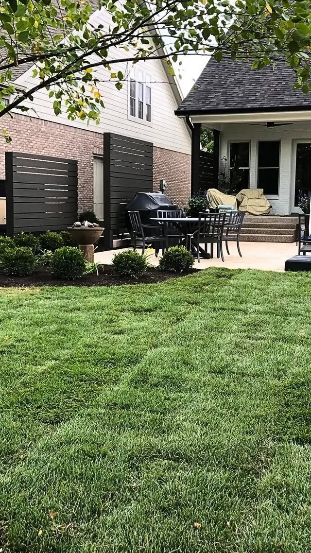 Front yard patio with black table and chairs beside a brick house and green lawn under trees