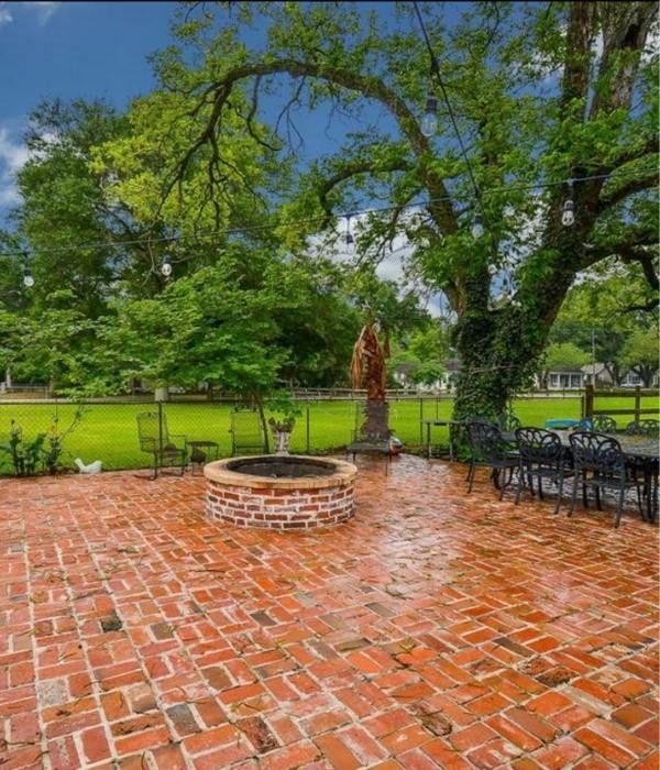 Brick patio with a circular fire pit, trees, and metal chairs overlooking a green lawn