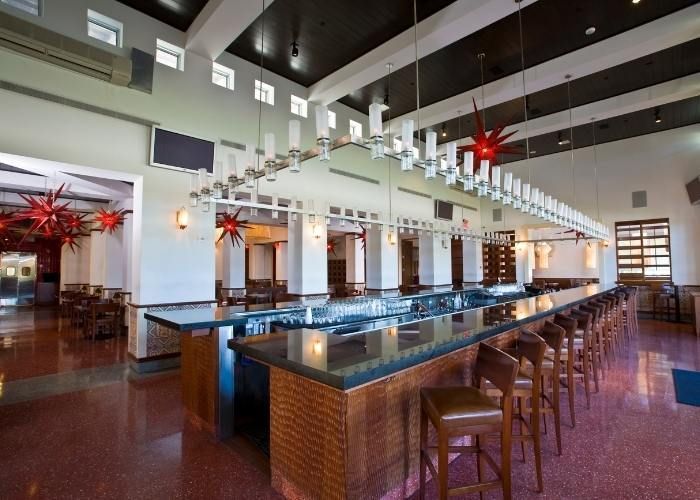 Modern bar interior with a long counter, stools, hanging lights, and wooden beams
