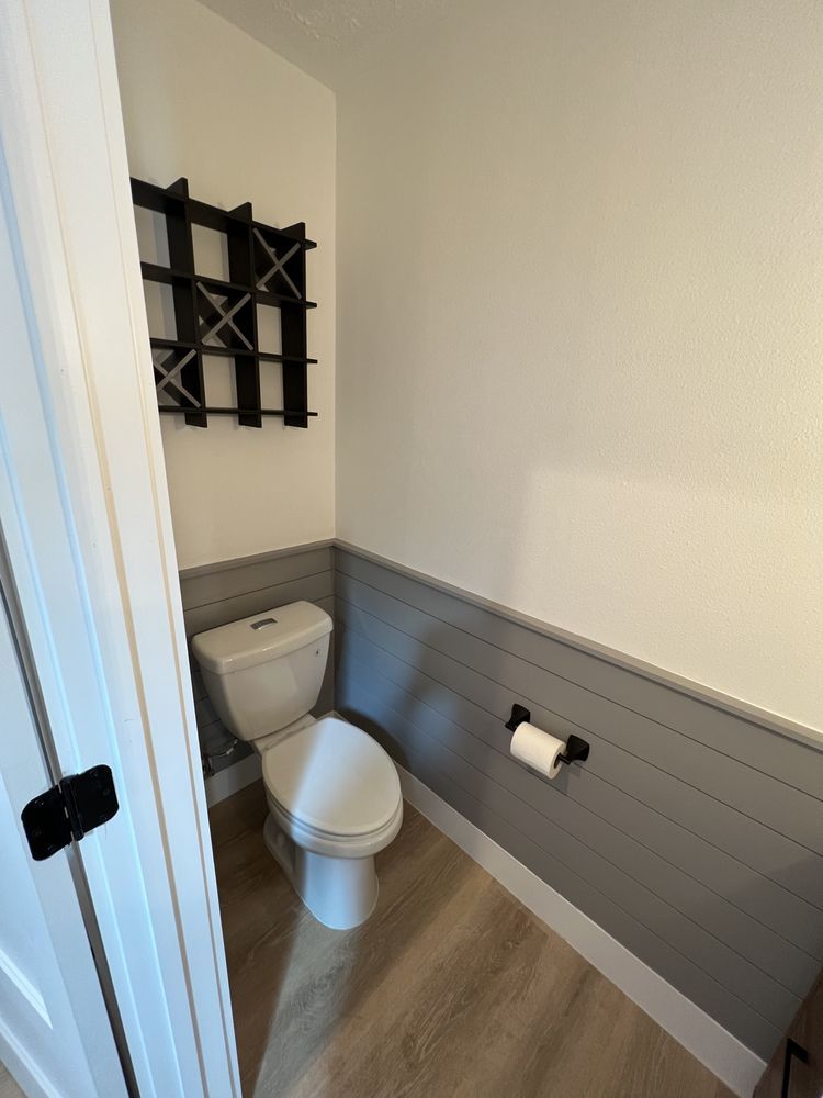 Small bathroom with white toilet, gray wainscoting, wood floor, and black wall shelf.