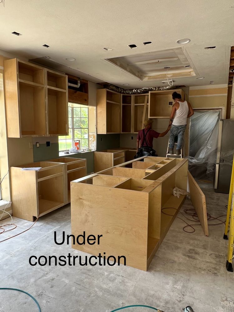 Kitchen under construction with unfinished wood cabinets, countertops, and two workers installing cabinets.