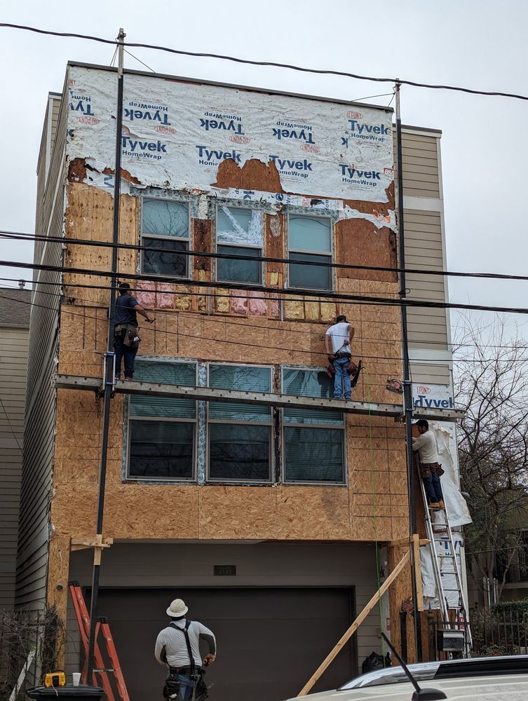 Workers on scaffolding repairing a brick building façade under plywood and window coverings.