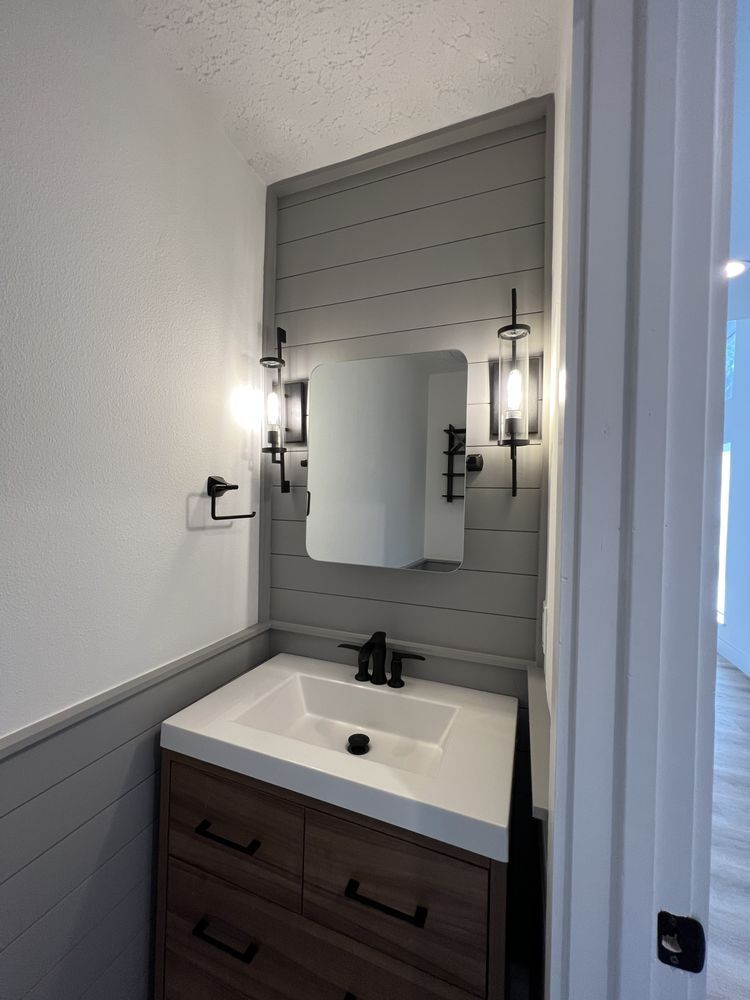 Small bathroom vanity with mirror, black faucet, sconce lights, and gray paneled wall.