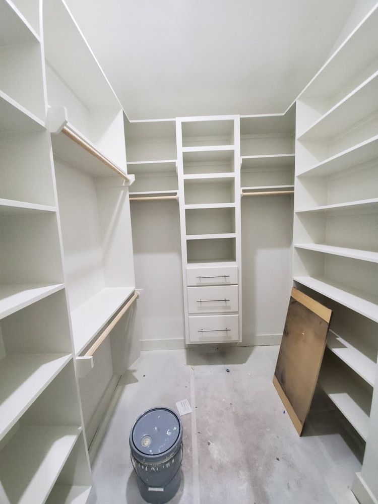 Empty white walk-in closet with built-in shelves, drawers, and a paint bucket on the floor