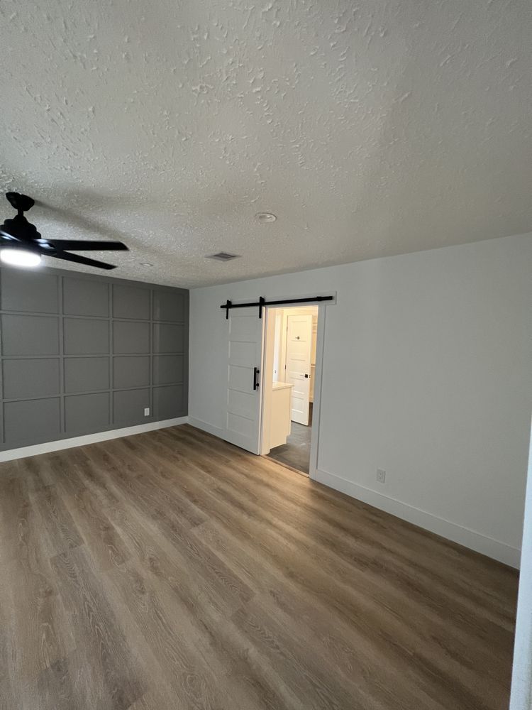 Empty modern bedroom with gray walls, wood-look floor, ceiling fan, and open doorway to a lit hall