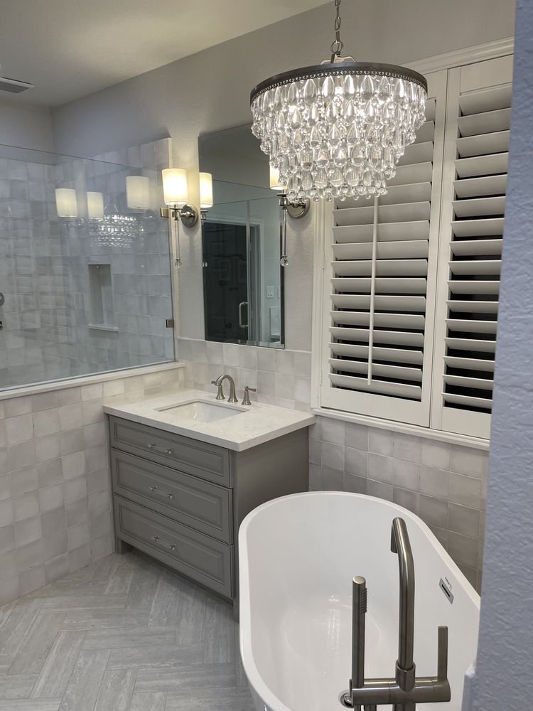 Modern bathroom with gray vanity, crystal chandelier, white shutters, and freestanding tub