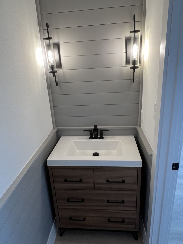 Small bathroom vanity with white sink, dark wood drawers, and wall sconces under a gray shiplap wall.