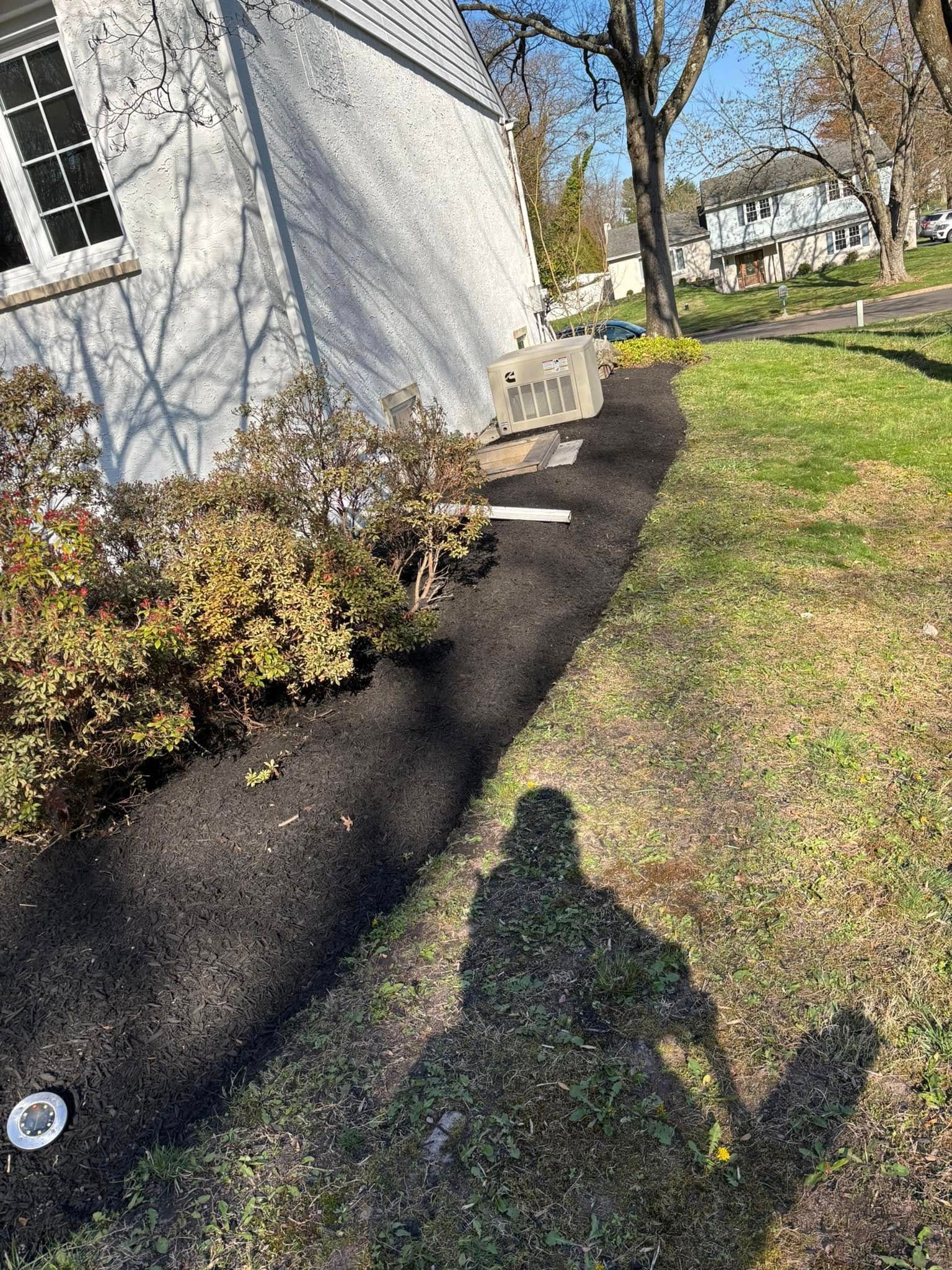 A person's shadow on grass next to a house with a dark mulch flower bed.