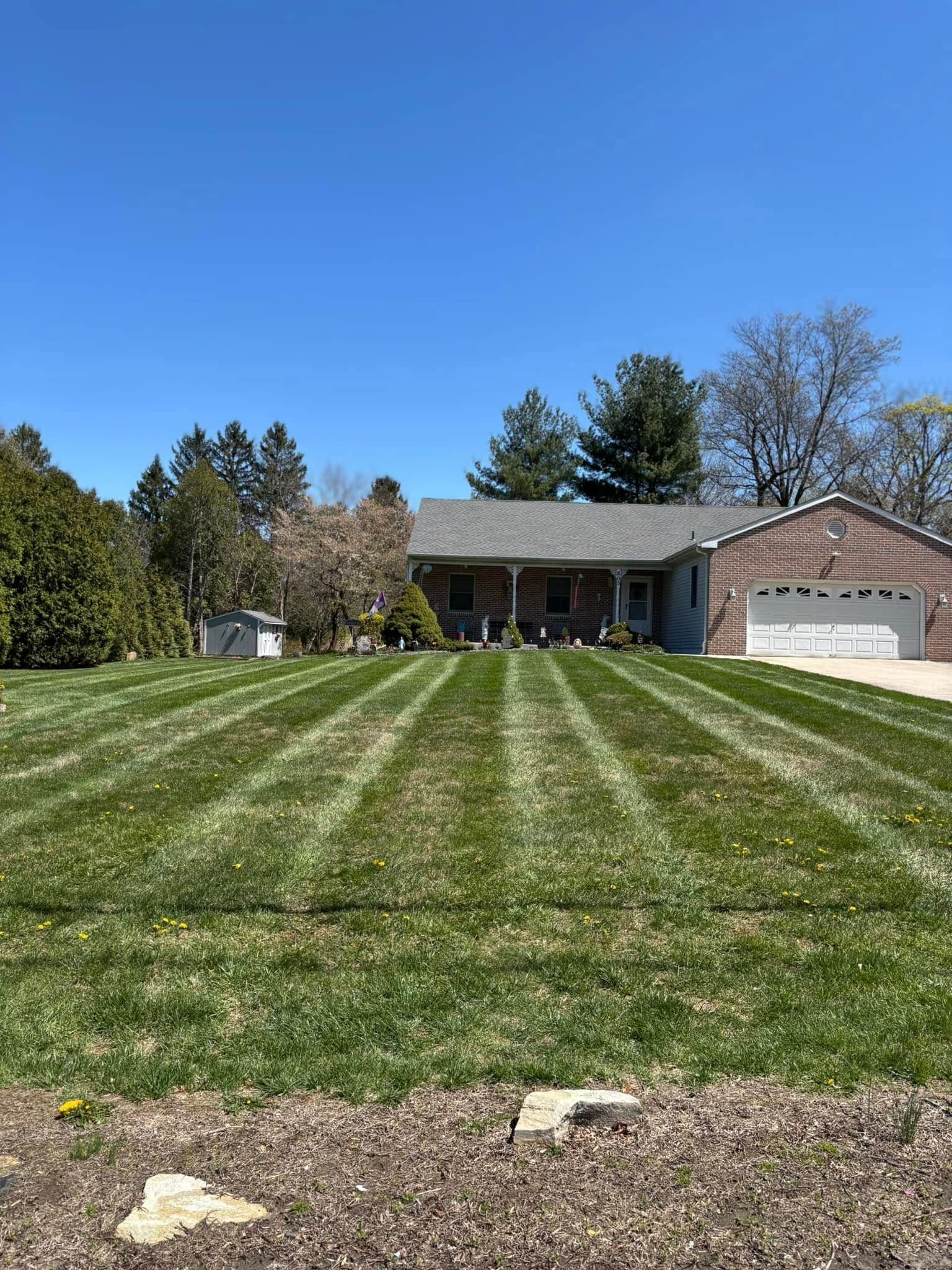 Green lawn with striped mowing pattern in front of a gray house with a porch and garage under a blue sky.