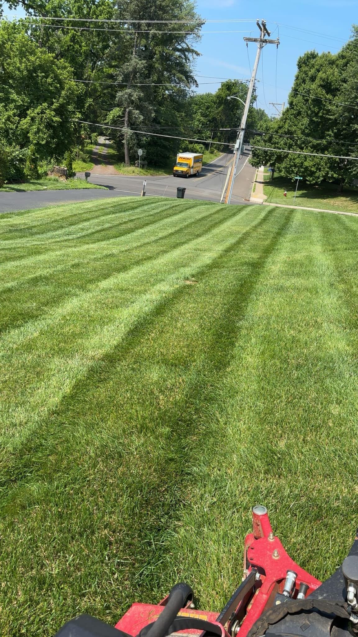 Lawn mower cutting grass on a sloped yard, with stripes. Yellow vehicle and power lines in the background.