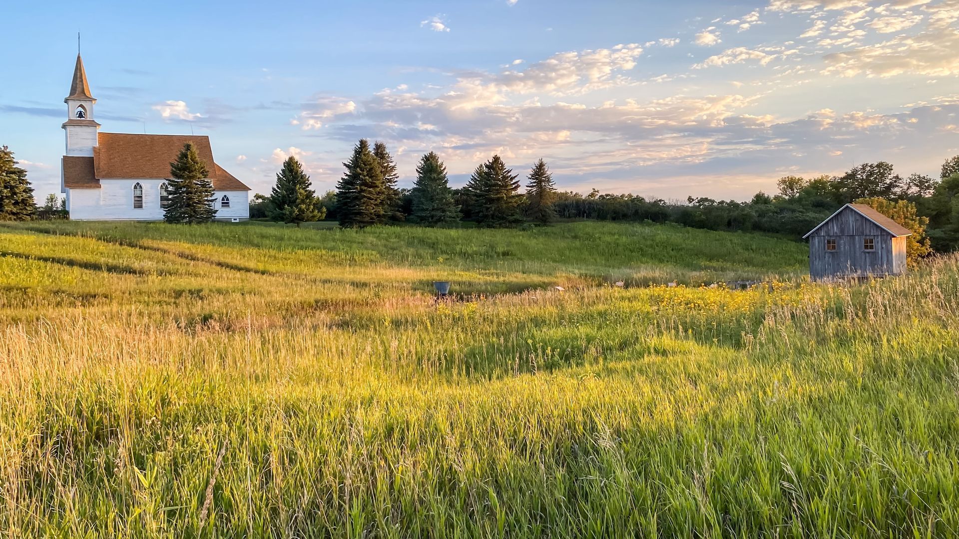 Sunlit church in a grassy field at sunset with a small shed and cloudy sky