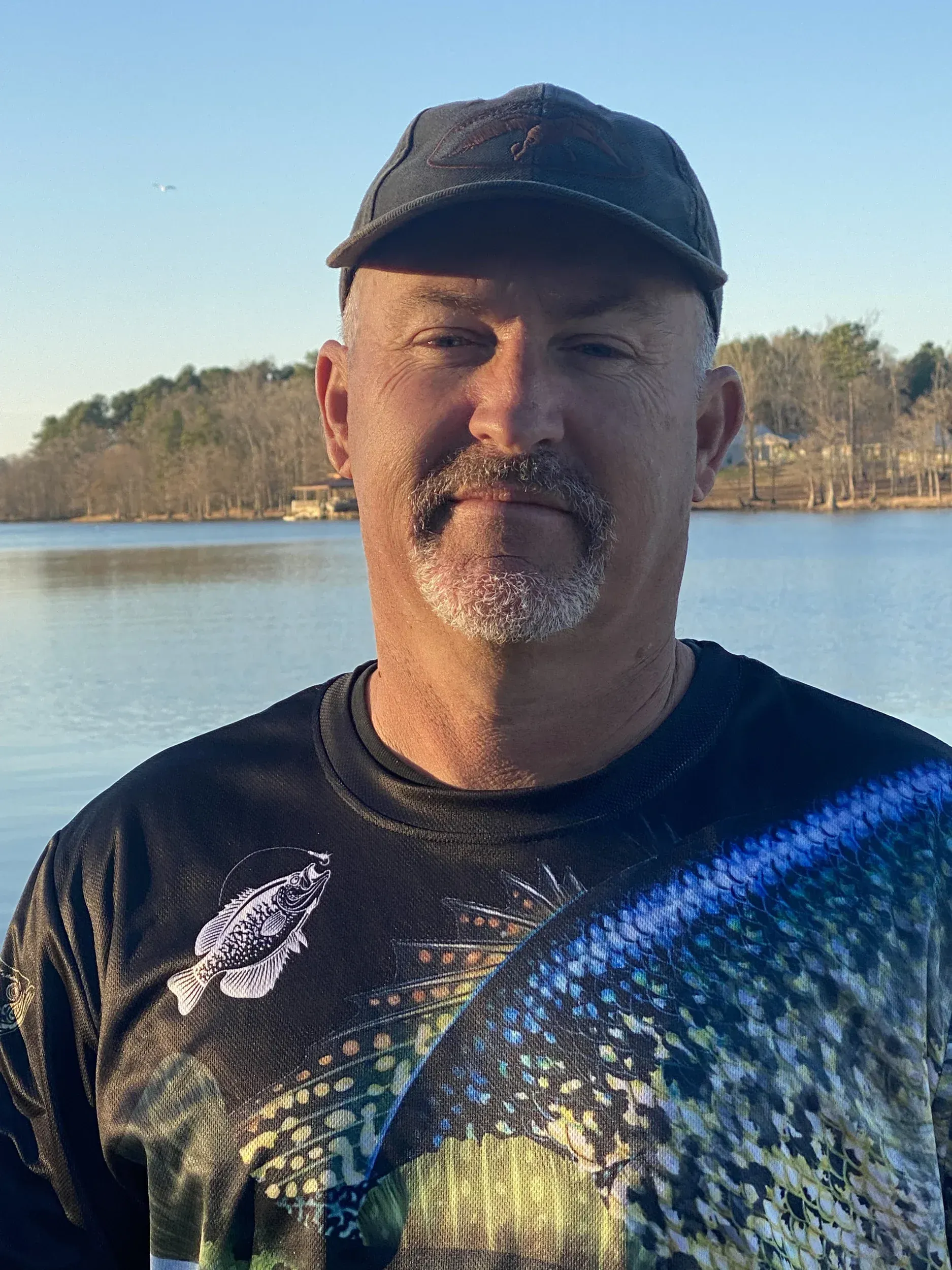 a man wearing a hat and a shirt with a fish on it is standing in front of a lake .