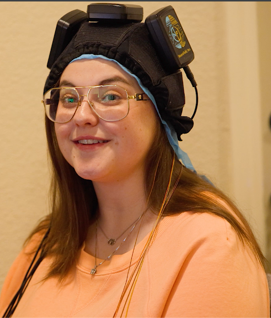 Smiling person wearing glasses and a black headpiece, seated indoors in a peach shirt.