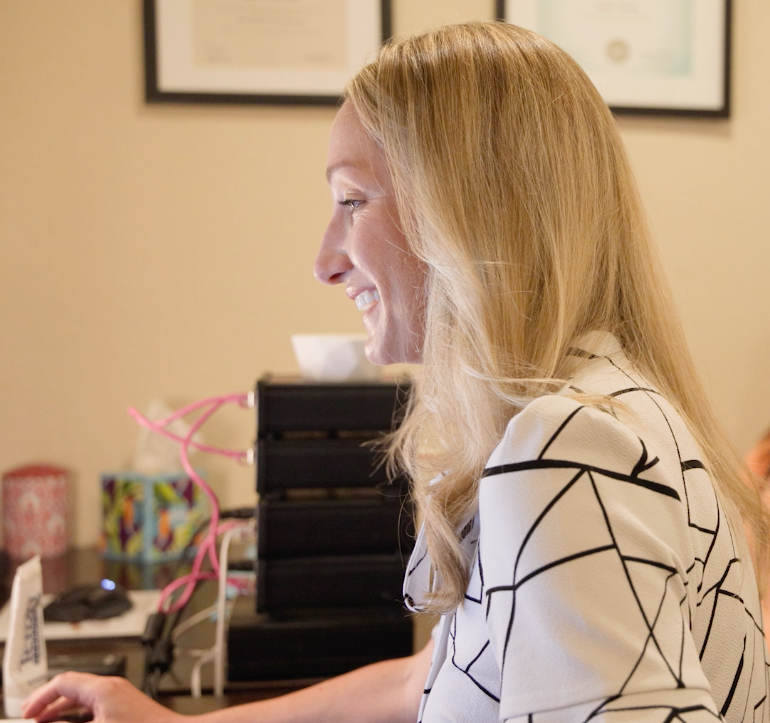 Woman smiling at a desk in an office, typing on a computer with framed art on the wall