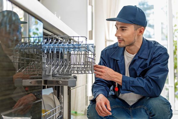 Technician inspecting a commercial dishwasher in a kitchen, wearing a blue cap and jacket.