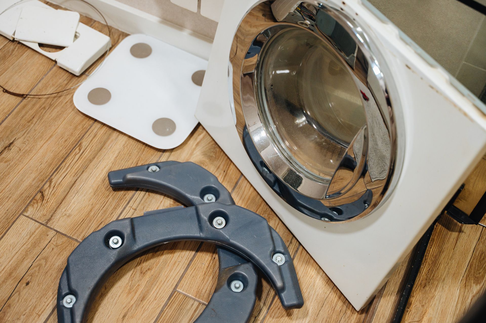 Broken washing machine on wooden floor with detached black plastic door ring and white parts nearby