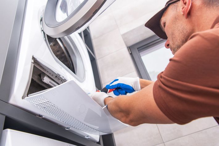 Technician cleaning a front-loading dryer with a spray bottle and cloth in a laundry room