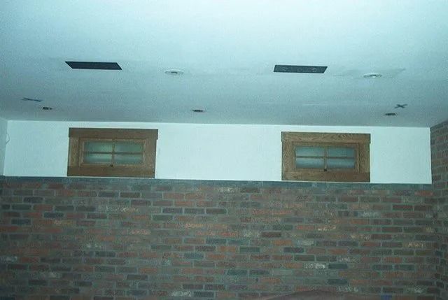 Brick wall with two small windows beneath a pale ceiling with vents.
