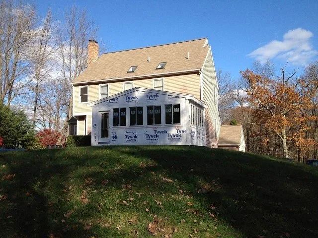 Two-story beige house with a white enclosed porch on a grassy hill under a blue sky