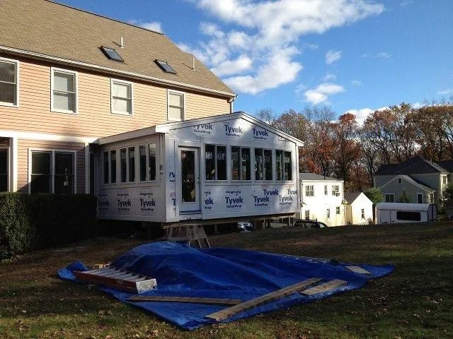 A white sunroom addition under construction beside a beige house, with blue tarp covering materials in the yard.