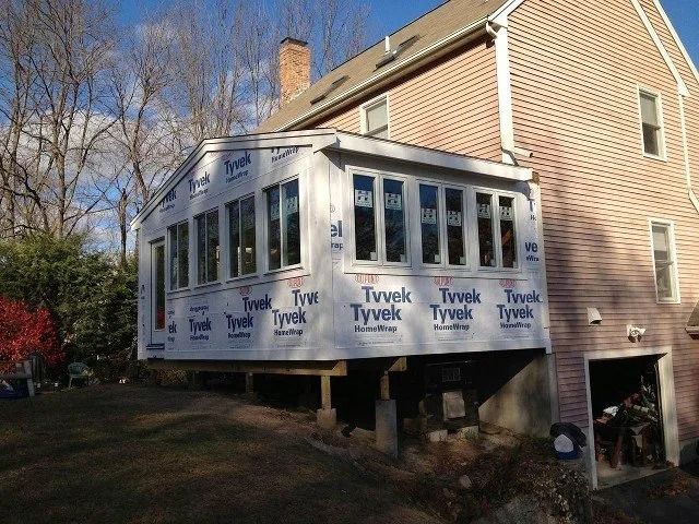 House addition under construction with white Tyvek wrap and many windows beside a brick home