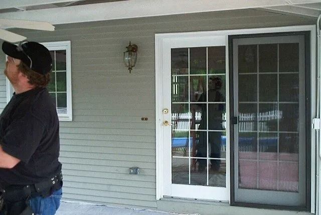 Man standing on a porch beside a sliding glass door and window with white trim