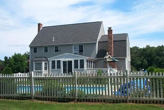 Gray two-story house with white trim, fenced yard, and a pool in front under a cloudy sky